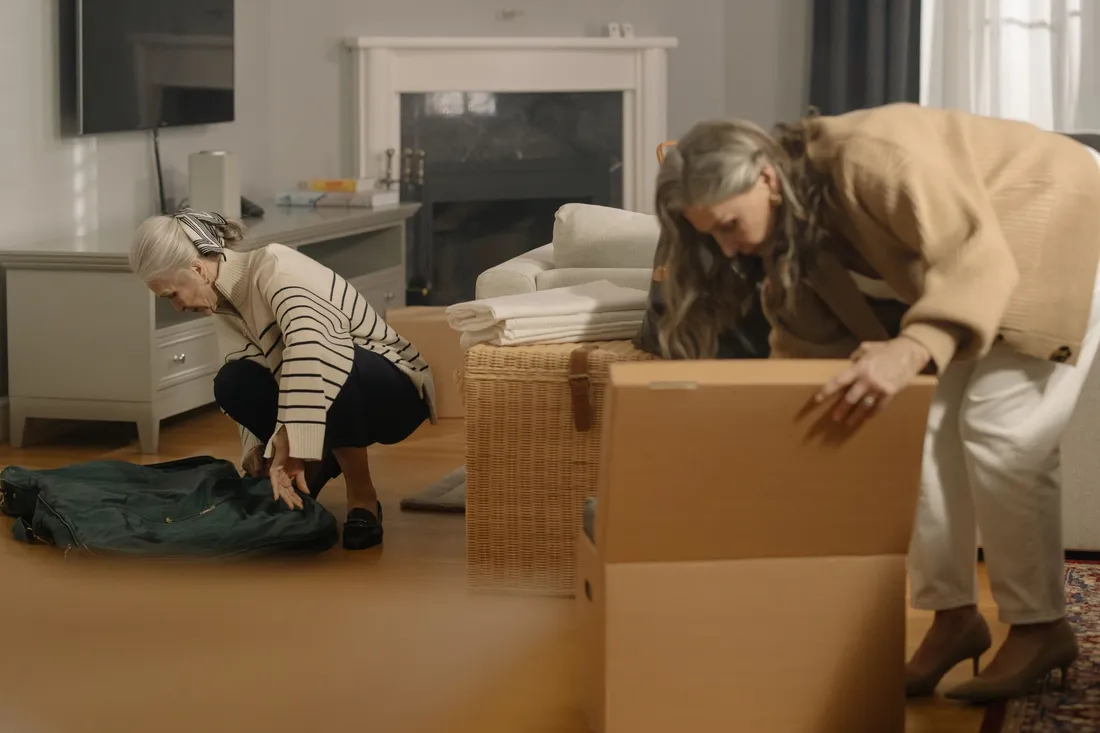Zwei ältere Frauen packen im Wohnzimmer Wäsche in einen Karton und eine Korbtruhe